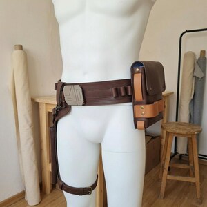 May include: A brown leather utility belt featuring a metallic geometric buckle, a large rectangular pouch with light tan leather accents, and a drop-leg holster strap. The belt includes integrated loops and is worn around the waist.
