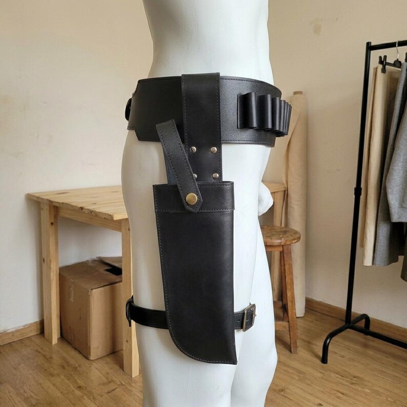 May include: A black leather belt featuring a wide waistband with integrated loops for small cylindrical items and a vertical holster attachment secured with a brass-colored metal snap button, worn around the waist and upper thigh with adjustable straps.