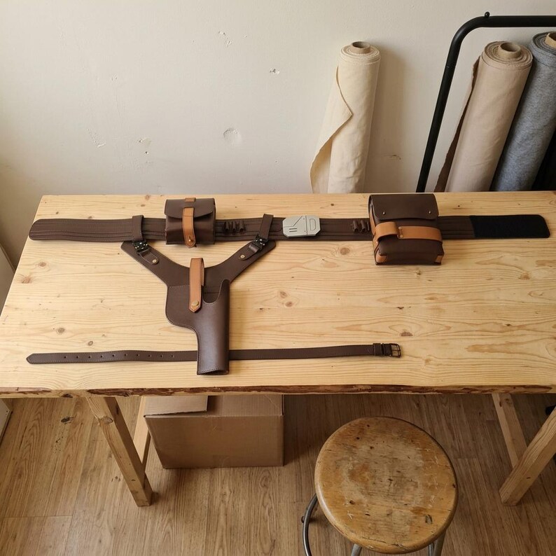 May include: A brown leather utility belt set featuring a holster, two pouches with tan straps, and a central silver-colored buckle. The set includes a Y-shaped harness attachment, a secondary thin strap, and integrated loops for small items.