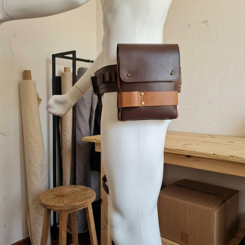May include: A dark brown leather hip bag with a rectangular flap and a horizontal tan leather strap across the front. The bag is attached to a wide, matching dark brown leather belt featuring multiple small loops along the side, secured with brass-colored metal rivets and fasteners.