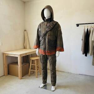 Pu&ograve; includere: Una giacca con cappuccio e pantaloni abbinati in una finitura marrone invecchiato con fasce arancioni sui polsini e sull'orlo. La giacca presenta una cerniera frontale, tasche multiple e una tracolla utilitaria marrone scuro.