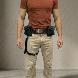 Op de afbeelding: Een roestkleurig T-shirt met korte mouwen gecombineerd met een beige broek en een zwarte utility-riem. De riem is voorzien van twee zwarte tasjes aan de zijkanten, een zilverkleurige rechthoekige gesp en een zwarte beenband.