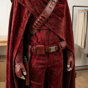 May include: A deep red textured cloak worn over a matching red shirt and trousers. A brown leather utility belt with a holster and pouches is worn at the waist, complemented by a matching brown leather bandolier strap across the chest and gloves.