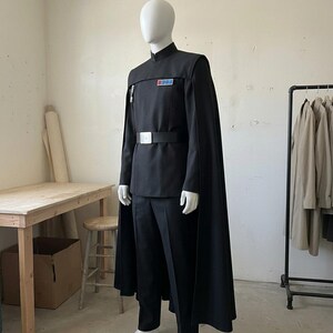 K&ouml;nnte beinhalten: Eine schwarze Uniform im Milit&auml;rstil, bestehend aus einer Tunika mit hohem Kragen, passender Hose und einem langen Umhang. Die Tunika verf&uuml;gt &uuml;ber ein rechteckiges Rangabzeichen mit blauen und roten Quadraten auf der Brust und einen breiten G&uuml;rtel mit silberner Schnalle.