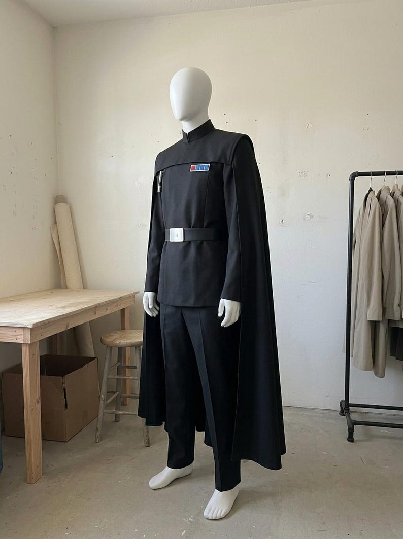 K&ouml;nnte beinhalten: Eine schwarze Uniform im Milit&auml;rstil, bestehend aus einer Tunika mit hohem Kragen, passender Hose und einem langen Umhang. Die Tunika verf&uuml;gt &uuml;ber ein rechteckiges Rangabzeichen mit blauen und roten Quadraten auf der Brust und einen breiten G&uuml;rtel mit silberner Schnalle.