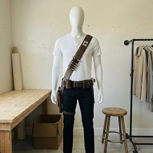 Pode incluir: Uma camisa henley branca, cal&ccedil;as de ganga azul-escuras e um cinto utilit&aacute;rio de couro castanho com al&ccedil;a de ombro a condizer. O cinto apresenta v&aacute;rios bolsos, um coldre para arma e uma s&eacute;rie de detalhes cil&iacute;ndricos met&aacute;licos na al&ccedil;a.