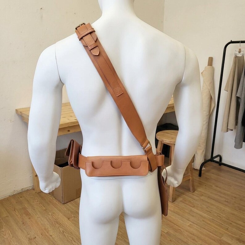May include: A tan leather utility belt and shoulder strap system worn across the torso. The belt features a horizontal pouch with three semi-circular cutouts for tool storage, a side pouch, and a long vertical holster attachment on the left side.