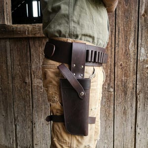 May include: A dark brown leather gun holster attached to a wide matching belt with integrated cartridge loops. The holster features a vertical strap with a metal snap closure and a secondary horizontal leg strap, secured with a metal buckle.