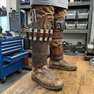 May include: Brown leather boots with a weathered appearance, featuring a zipper closure and a high-top design. The boots are part of a costume, with a holster and ammunition belt attached to the leg. The costume also includes brown pants.