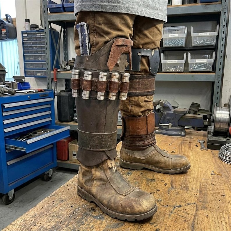 May include: Brown leather boots with a weathered appearance, featuring a zipper closure and a high-top design. The boots are part of a costume, with a holster and ammunition belt attached to the leg. The costume also includes brown pants.