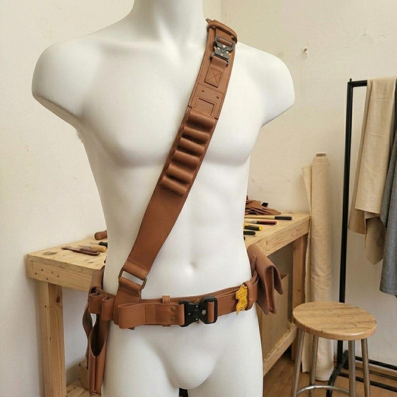 May include: A tan leather bandolier and utility belt set worn across the torso and waist. The gear features a wide shoulder strap with integrated loops, a waist belt with a black metal quick-release buckle, and attached leather pouches.