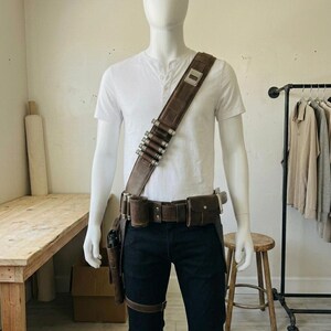 Pode incluir: Uma camisa henley branca e cal&ccedil;as escuras usadas com uma bandoleira de couro castanho com detalhes cil&iacute;ndricos prateados, um cinto utilit&aacute;rio a condizer com bolsas e um coldre preso &agrave; coxa num manequim sem rosto.