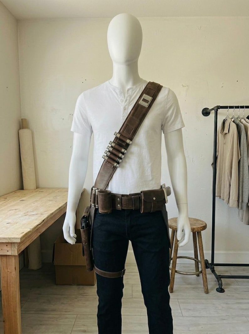 Pode incluir: Uma camisa henley branca e cal&ccedil;as escuras usadas com uma bandoleira de couro castanho com detalhes cil&iacute;ndricos prateados, um cinto utilit&aacute;rio a condizer com bolsas e um coldre preso &agrave; coxa num manequim sem rosto.