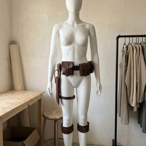 May include: A brown leather utility belt set featuring multiple pouches, a holster attached to a thigh strap, and two matching leg bands. The set is worn around the waist and legs, constructed with dark brown leather and secured with metal hardware.