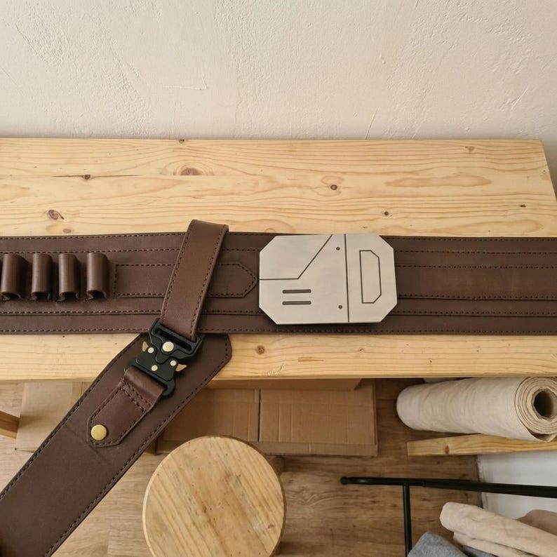 May include: A brown utility belt featuring a series of loops for holding small items, a metallic silver rectangular buckle with geometric engravings, and a black tactical-style quick-release buckle attached to a secondary strap section.