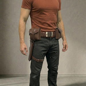 Op de afbeelding: Een roestkleurig T-shirt met korte mouwen gecombineerd met een donkergrijze broek en een bruine leren utility-riem. De riem heeft een grote rechthoekige zilveren gesp, twee zijzakken en een lang holster dat met een beenband is bevestigd.
