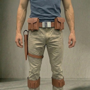 May include: A brown leather utility belt system featuring two rectangular pouches, a large silver-toned buckle, and a holster attached to a thigh strap. The set includes two matching brown leather leg bands with integrated loops, worn over tan trousers.