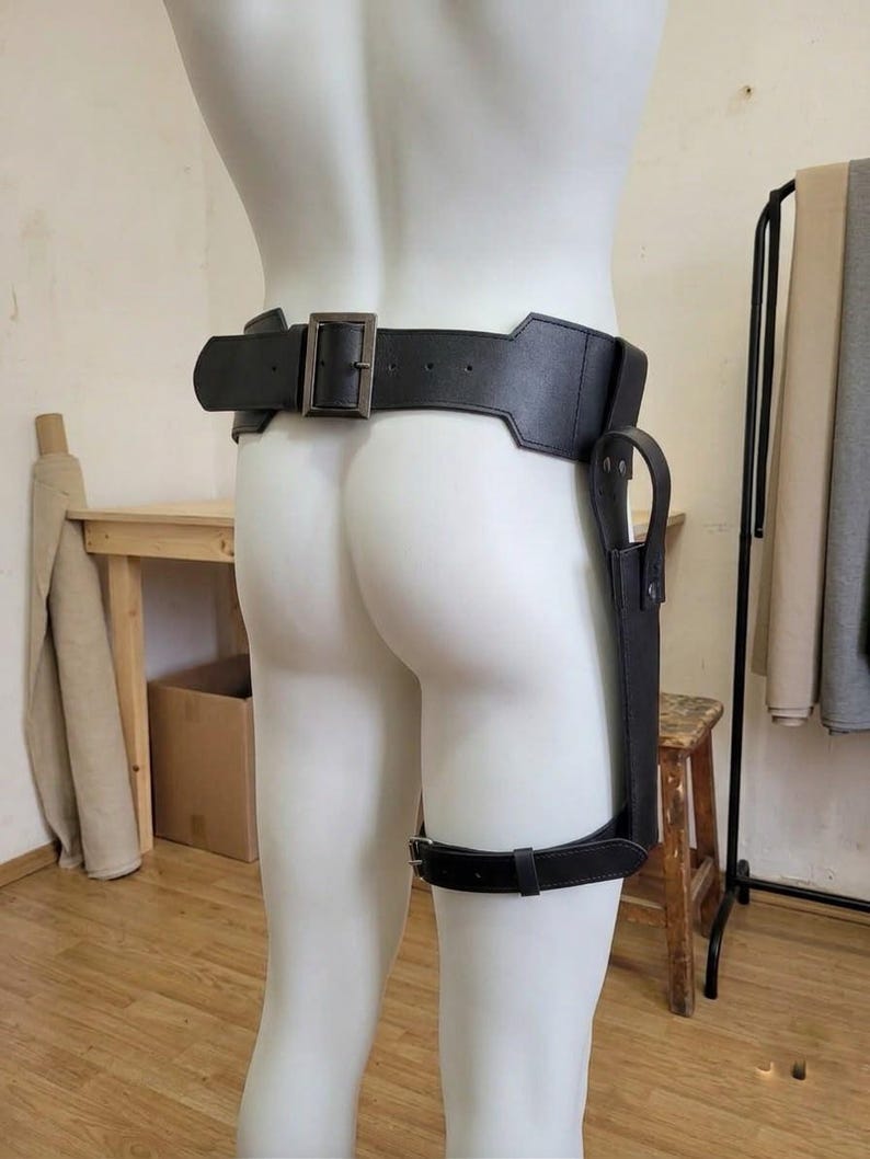 May include: A black leather-like belt featuring a wide waist band with a rectangular metal buckle and an integrated holster attachment that extends down to a secondary adjustable leg strap, designed to be worn around the waist and thigh.