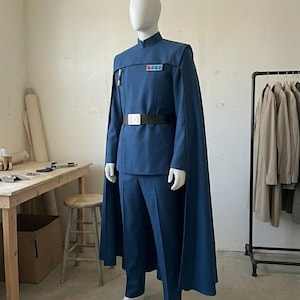 K&ouml;nnte beinhalten: Ein blaues Uniformset bestehend aus einer Tunika mit Stehkragen und Umhang, passender Hose und einem schwarzen G&uuml;rtel mit silberner Schnalle. Die Tunika hat ein rechteckiges Rangabzeichen mit farbigen Quadraten auf der Brust und einen Zylinder.