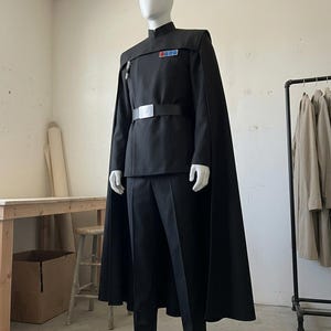 K&ouml;nnte beinhalten: Eine schwarze Uniform bestehend aus einer lang&auml;rmeligen Tunika mit Stehkragen, einer passenden Hose und einem bodenlangen Umhang. Die Tunika verf&uuml;gt &uuml;ber ein rechteckiges Rangabzeichen mit farbigen Quadraten auf der Brust und einen schwarzen G&uuml;rtel.