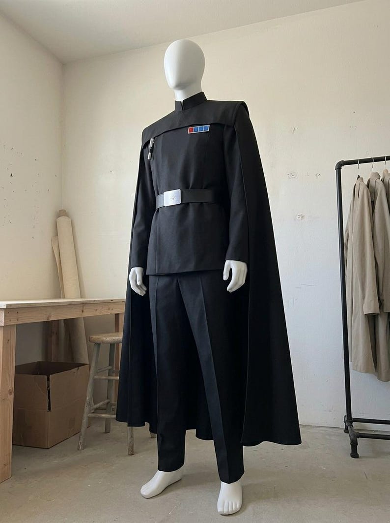 K&ouml;nnte beinhalten: Eine schwarze Uniform bestehend aus einer lang&auml;rmeligen Tunika mit Stehkragen, einer passenden Hose und einem bodenlangen Umhang. Die Tunika verf&uuml;gt &uuml;ber ein rechteckiges Rangabzeichen mit farbigen Quadraten auf der Brust und einen schwarzen G&uuml;rtel.