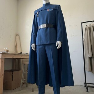K&ouml;nnte beinhalten: Ein blaues Uniformset im Milit&auml;rstil, bestehend aus einer lang&auml;rmeligen Tunika mit Stehkragen, einer passenden Hose und einem bodenlangen Umhang. Die Tunika verf&uuml;gt &uuml;ber ein rechteckiges Rangabzeichen mit farbigen Quadraten.