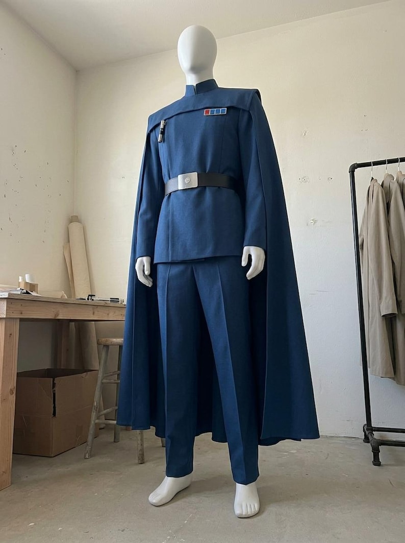 K&ouml;nnte beinhalten: Ein blaues Uniformset im Milit&auml;rstil, bestehend aus einer lang&auml;rmeligen Tunika mit Stehkragen, einer passenden Hose und einem bodenlangen Umhang. Die Tunika verf&uuml;gt &uuml;ber ein rechteckiges Rangabzeichen mit farbigen Quadraten.
