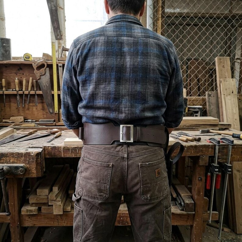May include: A wide, dark brown leather tool belt with a silver-toned metal buckle worn over brown canvas work pants. The belt features a sturdy construction with visible stitching, designed to sit securely around the waist for carrying various tools.