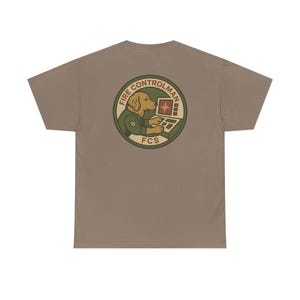 May include: A tan t-shirt featuring a circular graphic of a golden retriever in a military uniform operating a computer. The graphic reads "FIRE CONTROLMAN" and "FCS" in a circular design with olive green and brown accents.