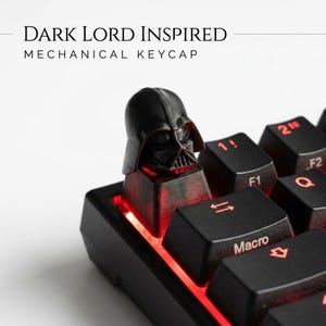 May include: A black mechanical keycap shaped like a helmeted head, positioned on a keyboard with red backlighting. The text "DARK LORD INSPIRED MECHANICAL KEYCAP" is visible above the keyboard. The keycap features a detailed mask design.