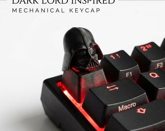 Puede incluir: Una tecla mecánica negra con forma de cabeza con casco, situada en un teclado con retroiluminación roja. El texto "DARK LORD INSPIRED MECHANICAL KEYCAP" es visible arriba. La tecla presenta un diseño de máscara muy detallado.