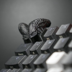 May include: A black Alien-themed keycap set on a computer keyboard. The keycap features a detailed, textured design of the iconic creature. The keyboard keys are black and gray, with white lettering.
