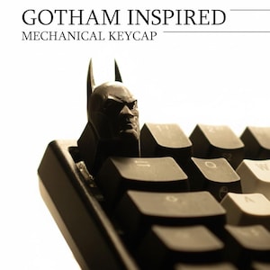 May include: A black mechanical keycap featuring a Batman head design, set against a keyboard. The keycap is a detailed sculpture of the superhero's face, with the text "Gotham Inspired Mechanical Keycap" above.
