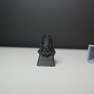 May include: A black, textured sculpture of a head with a detailed surface, mounted on a rectangular base. A light purple, square-shaped object with a plus sign is to the right. The objects are on a white surface.