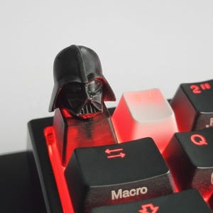 May include: A black Darth Vader keycap on a computer keyboard. The keycap is detailed with the character's helmet and face. The keyboard keys are black with red backlighting, and some keys have white lettering.