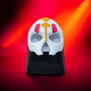 May include: A white skull-shaped keycap with red and gold detailing. The keycap is mounted on a black base, set against a red and orange gradient backdrop. The design features a stylized skull with eye sockets and a central vertical line.