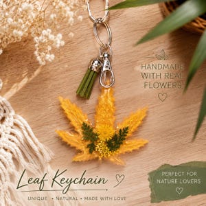 Peut inclure: Un porte-clés en résine jaune en forme de feuille de cannabis, incrusté de petites fougères vertes et de fleurs jaunes. Il comprend un fermoir mousqueton en métal argenté, un anneau et un pompon vert. Le texte indique : HANDMADE WITH REAL FLOWERS.