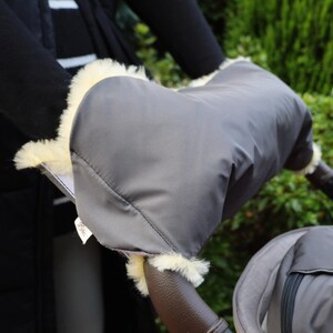 Könnte beinhalten: Grauer Kinderwagen-Handwärmer mit cremefarbenem Kunstfellbesatz. Der Handwärmer ist an einem braunen Leder-Kinderwagengriff befestigt. Das Bild zeigt eine Nahaufnahme des Kinderwagenzubehörs.