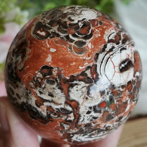 Peut inclure: Une sphère polie avec un motif unique de tourbillons rouges, noirs et blancs. La surface de l'orbite a un aspect marbré, avec des veines et des variations de couleurs complexes. La sphère est un objet décoratif.