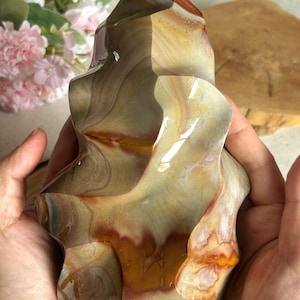 Pode incluir: Uma escultura de pedra polida em forma de chama com faixas onduladas em tons de bege, creme, vermelho escuro e castanho. A superfície é lisa e reflexiva, exibindo padrões minerais naturais com uma orientação vertical e curvas orgânicas.