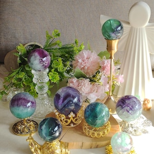 Peut inclure: Une collection de sphères en fluorine polie dans des tons de violet, vert et bleu sarcelle, présentées sur divers supports décoratifs, notamment des couronnes en métal doré, une figurine d'escargot doré et des supports en verre transparent.