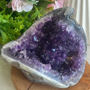 May include: A large amethyst geode with a deep purple crystal interior. The geode is set on a round, natural wood base. The outer edge of the geode is a mix of gray and white. Pink flowers and green foliage are in the background.