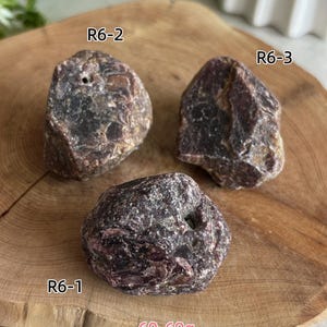 Peut inclure: Trois pierres brutes brun-rouge fonc&eacute; aux surfaces irr&eacute;guli&egrave;res et textur&eacute;es. Chaque pierre est &eacute;tiquet&eacute;e avec un code : R6-1, R6-2 et R6-3. Le texte 60-69g est visible en bas, indiquant la plage de poids de ces sp&eacute;cimens min&eacute;raux.