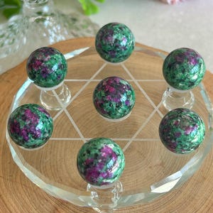 May include: Seven polished ruby in zoisite spheres arranged on a clear glass plate etched with a six-pointed star design. Each sphere features a mottled pattern of vibrant magenta-pink ruby inclusions contrasted against a deep green zoisite matrix.