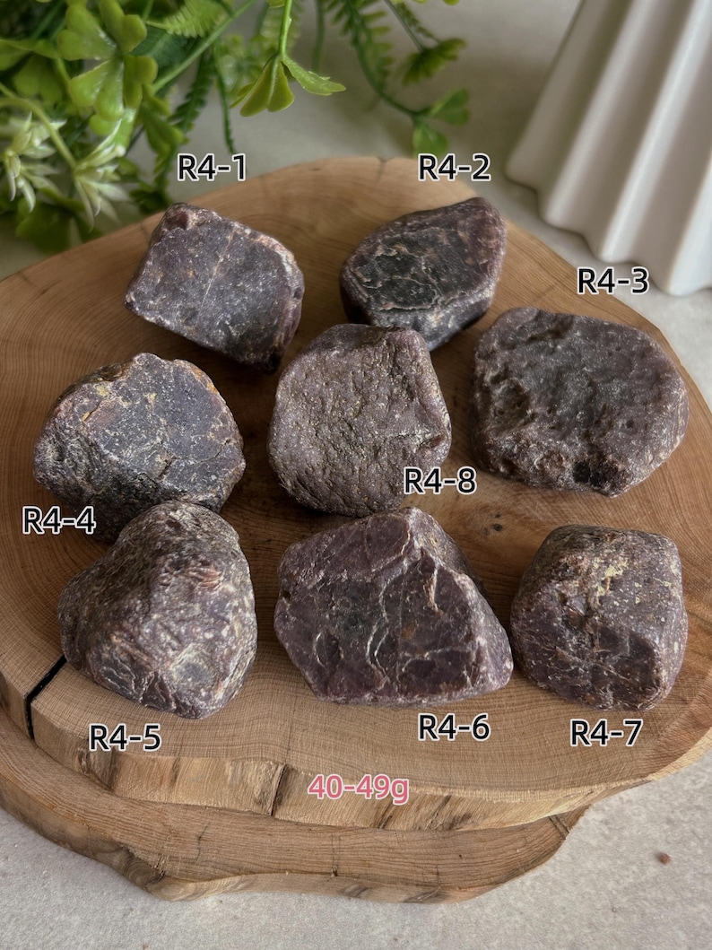 Peut inclure: Une collection de huit pierres brutes rouge-violet fonc&eacute; &eacute;tiquet&eacute;es R4-1 &agrave; R4-8, avec une plage de poids de 40-49g. Les pierres ont des surfaces irr&eacute;guli&egrave;res et textur&eacute;es et sont dispos&eacute;es sur une tranche de bois circulaire avec des grains.
