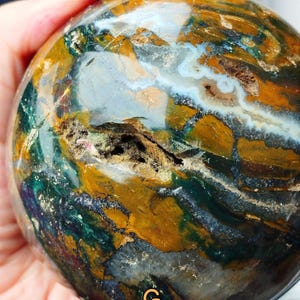 Puede incluir: Una esfera de piedra preciosa pulida con un patrón complejo de colores. La esfera presenta una mezcla de tonos verdes, marrones y blancos, con áreas de azul oscuro y dorado. La superficie tiene un aspecto natural y texturizado, con una pequeña cavidad.