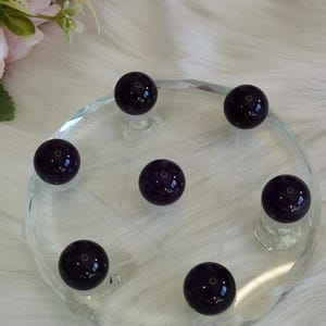 Puede incluir: Un conjunto de seis piedras de amatista esféricas, lisas y de color púrpura oscuro, dispuestas en un patrón circular sobre un plato de exhibición de vidrio transparente, circular y facetado. Cada piedra está pulida con brillo.