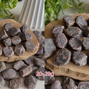 Peut inclure: Une collection de pierres de grenat brutes, brun rouge&acirc;tre fonc&eacute;, aux formes irr&eacute;guli&egrave;res et facett&eacute;es. Les pierres sont &eacute;parpill&eacute;es sur une surface en bois clair, avec une plage de poids de 20-29g imprim&eacute;e en texte rose sur l'image.