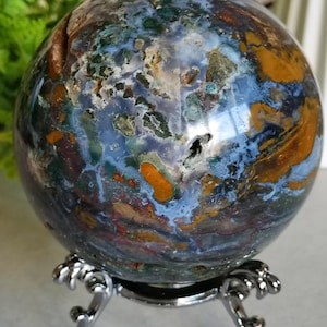 Puede incluir: Una esfera de piedra preciosa pulida con un patrón arremolinado de tonos azules, verdes, marrones y naranjas. Se exhibe sobre un soporte decorativo de metal plateado. Objeto decorativo.