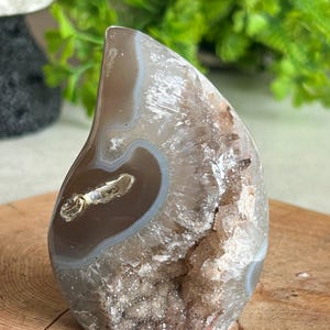 Puede incluir: Una piedra de ágata pulida con forma de lágrima, que presenta superficies lisas y bandeadas de color marrón y gris en un lado y una cavidad de geoda cristalina texturizada con formaciones minerales blancas y marrón claro brillantes.
